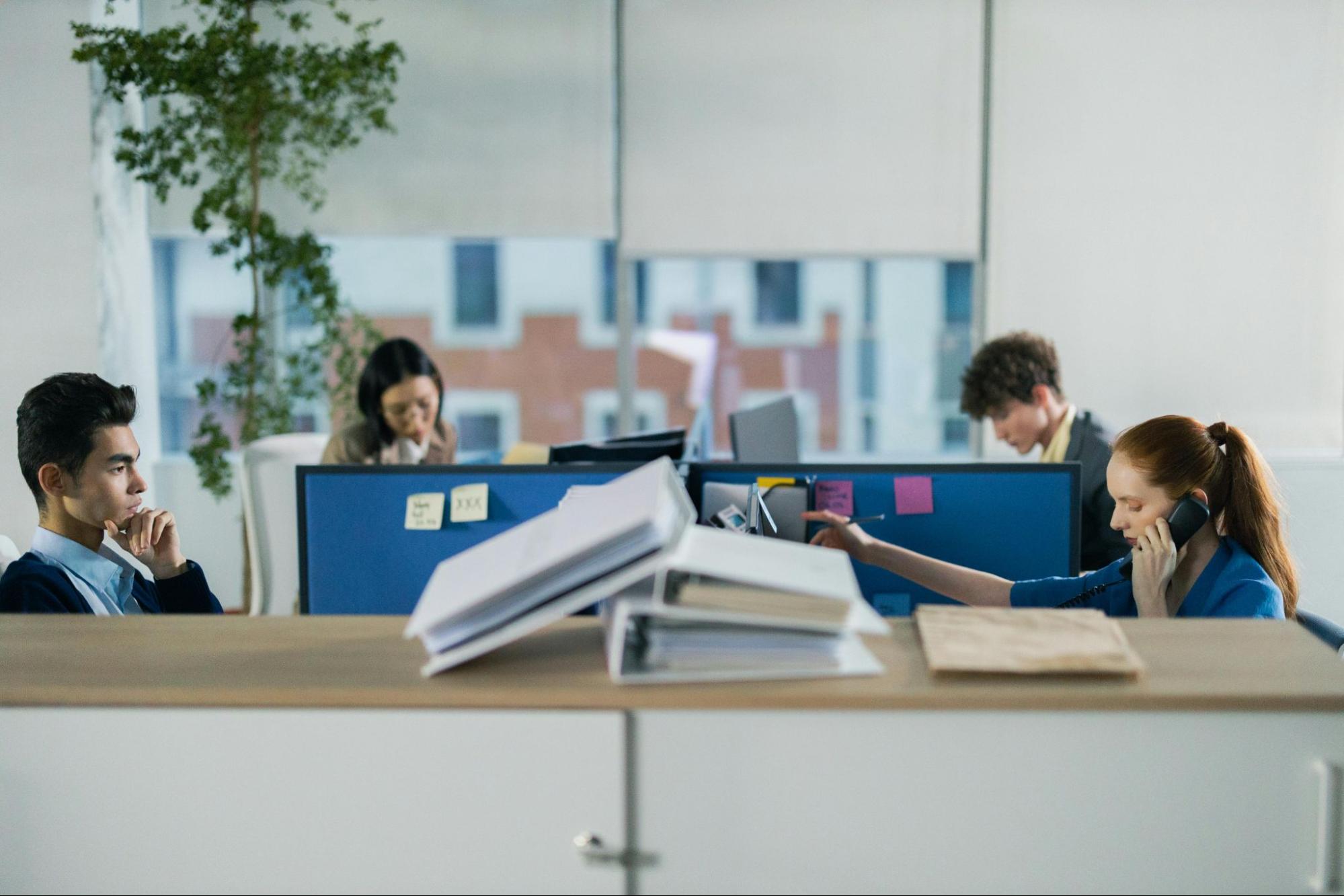 4 busy persons in cubical desk with 3 white books as foreground